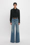 A person stands against a white background, embodying contemporary timeless fashion with the Victoria Beckham Asymmetric Ruffle Blouse In Black and light blue wide-legged jeans cinched with a belt.