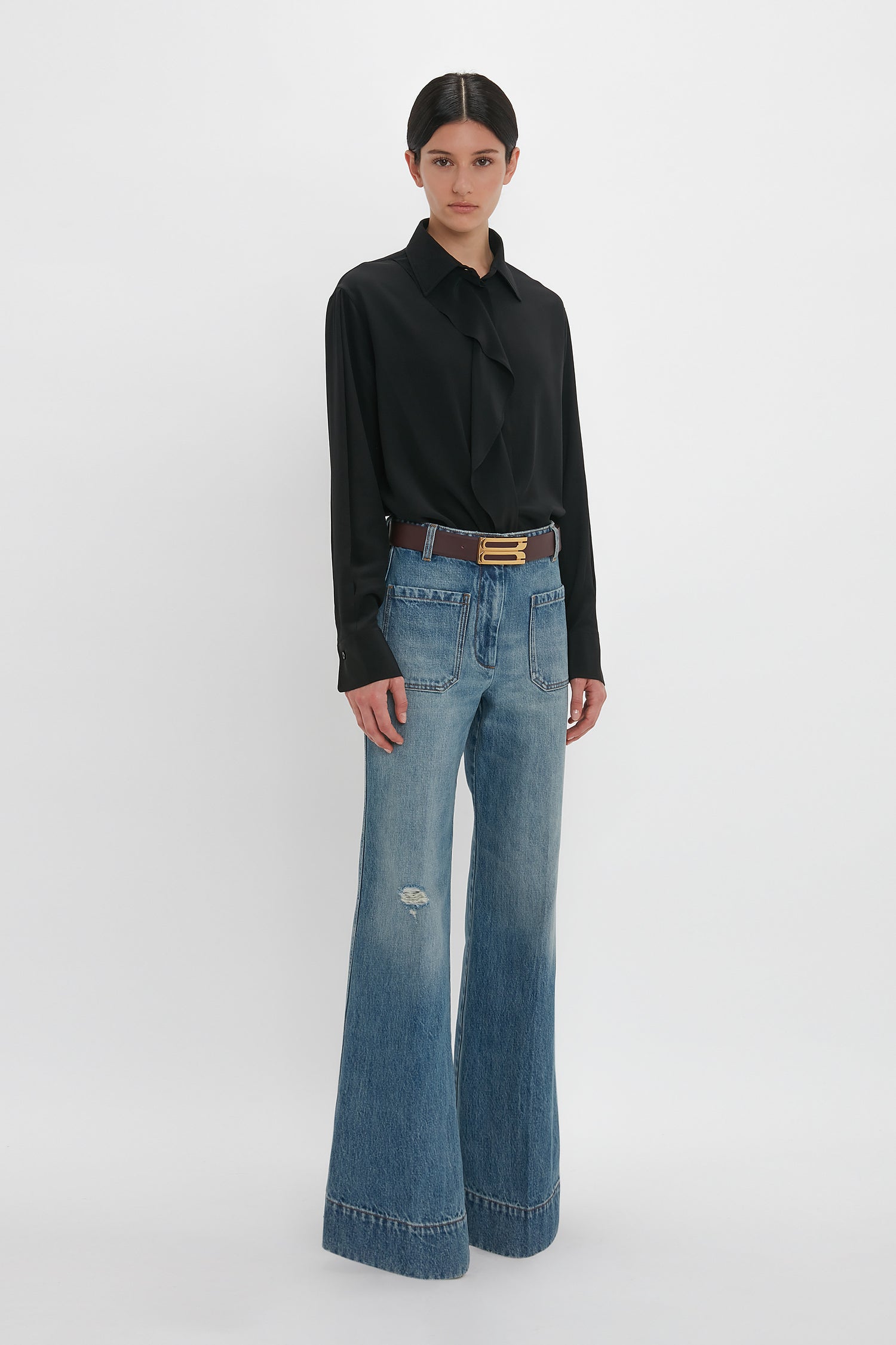 A person stands against a plain white background wearing a long-sleeved black blouse, wide-leg Alina High Waisted Jean In Worn Blue Wash by Victoria Beckham, and brown shoes. The jeans have a slightly frayed hem and a small rip on the left knee.