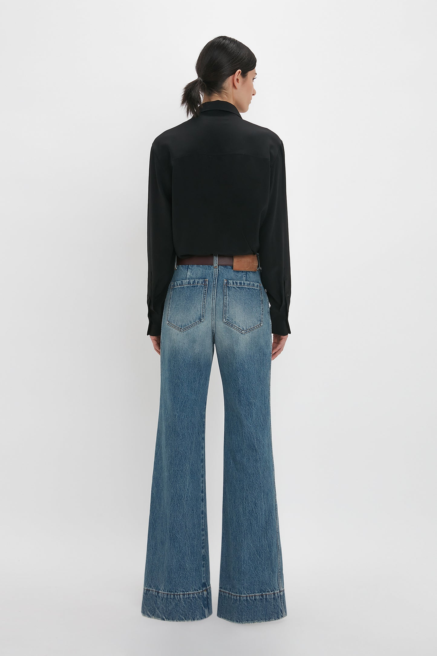 A person with long dark hair tied back is standing facing away, wearing a black long-sleeve shirt and high-waisted Alina High Waisted Jean In Worn Blue Wash from Victoria Beckham with frayed hems.