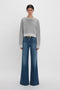 Woman standing in a studio, wearing a Victoria Beckham grey marl cropped sweatshirt and blue flared jeans with a thin belt.