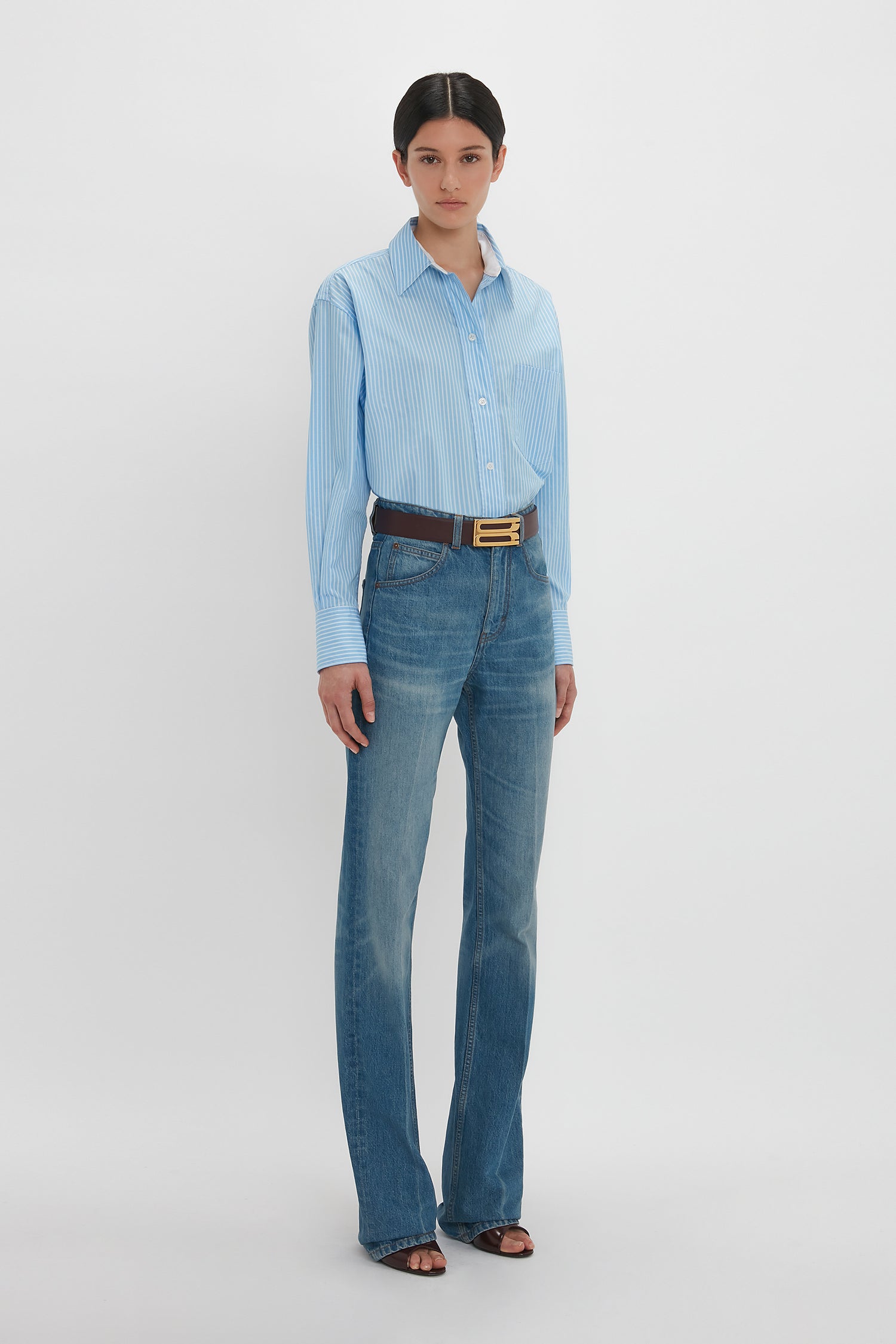 A person stands against a plain white background, wearing a Victoria Beckham Cropped Long Sleeve Shirt in Marina-White tucked into blue jeans with a brown belt. They are also wearing black open-toed shoes.