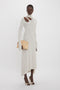 A person in a Long Sleeve Cut-Out Jersey Midi Dress In Bone by Victoria Beckham with an asymmetric hemline and a beige clutch, wearing white high-heeled shoes, poses against a plain white background.