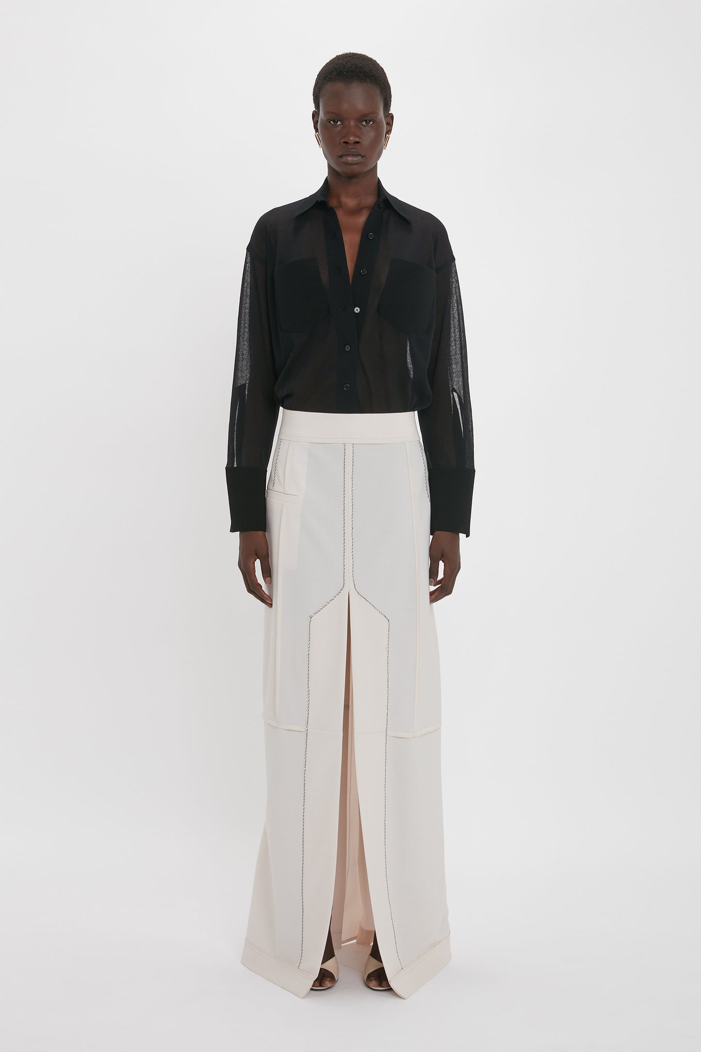 A person stands posing in a relaxed fit black Pocket Detail Shirt In Black by Victoria Beckham and a long, high-waisted white skirt with a central slit. The background is plain white.