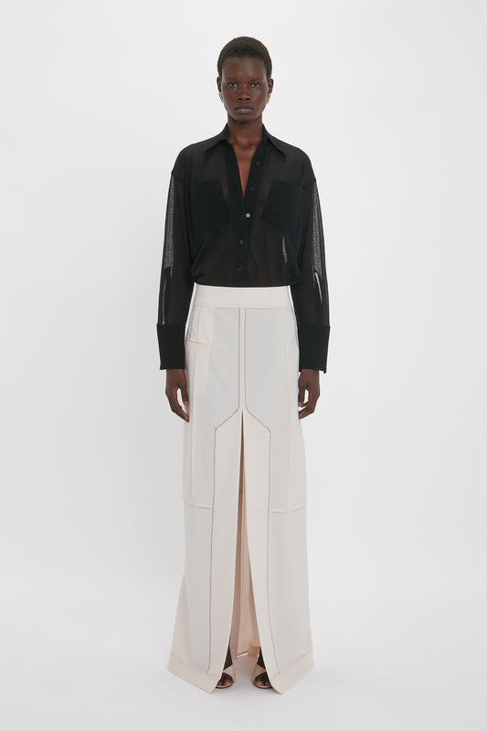 A person is standing against a white background, wearing a sheer black long-sleeve shirt and a high-waisted Victoria Beckham Deconstructed Floor-Length Skirt In Bone with a front slit and exposed seams.