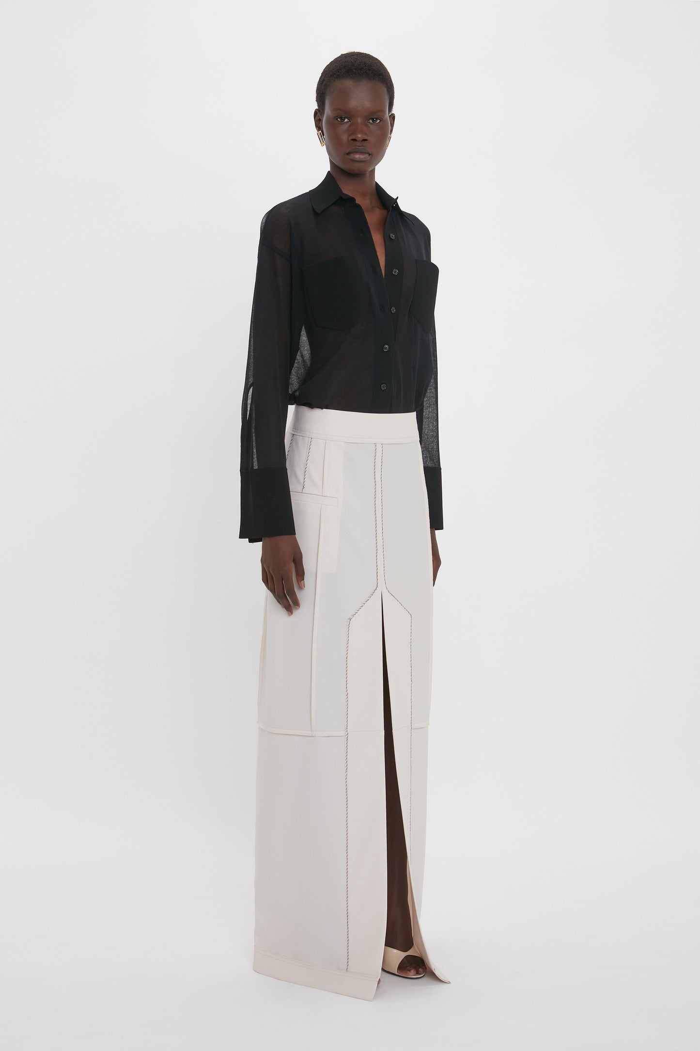 A person stands against a plain background wearing a sheer black Pocket Detail Shirt In Black by Victoria Beckham and a long off-white skirt with a central slit.