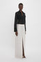 A person stands against a plain background wearing a sheer black Pocket Detail Shirt In Black by Victoria Beckham and a long off-white skirt with a central slit.