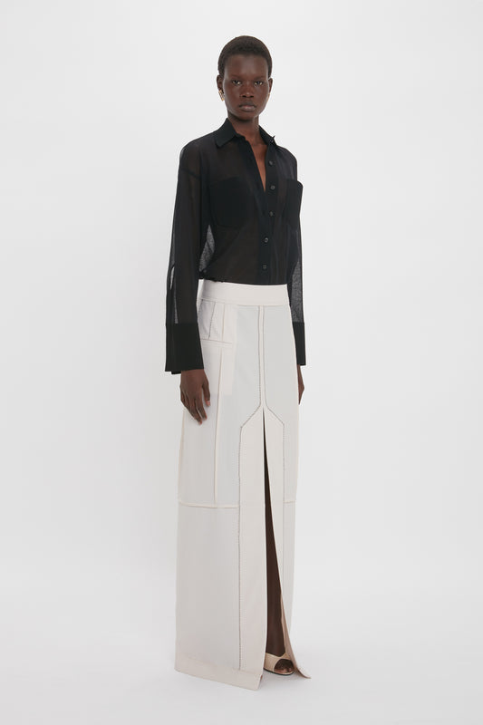 A person standing, wearing a black sheer long-sleeve button-up shirt and a Victoria Beckham Deconstructed Floor-Length Skirt In Bone with a front slit and box pleat detail.