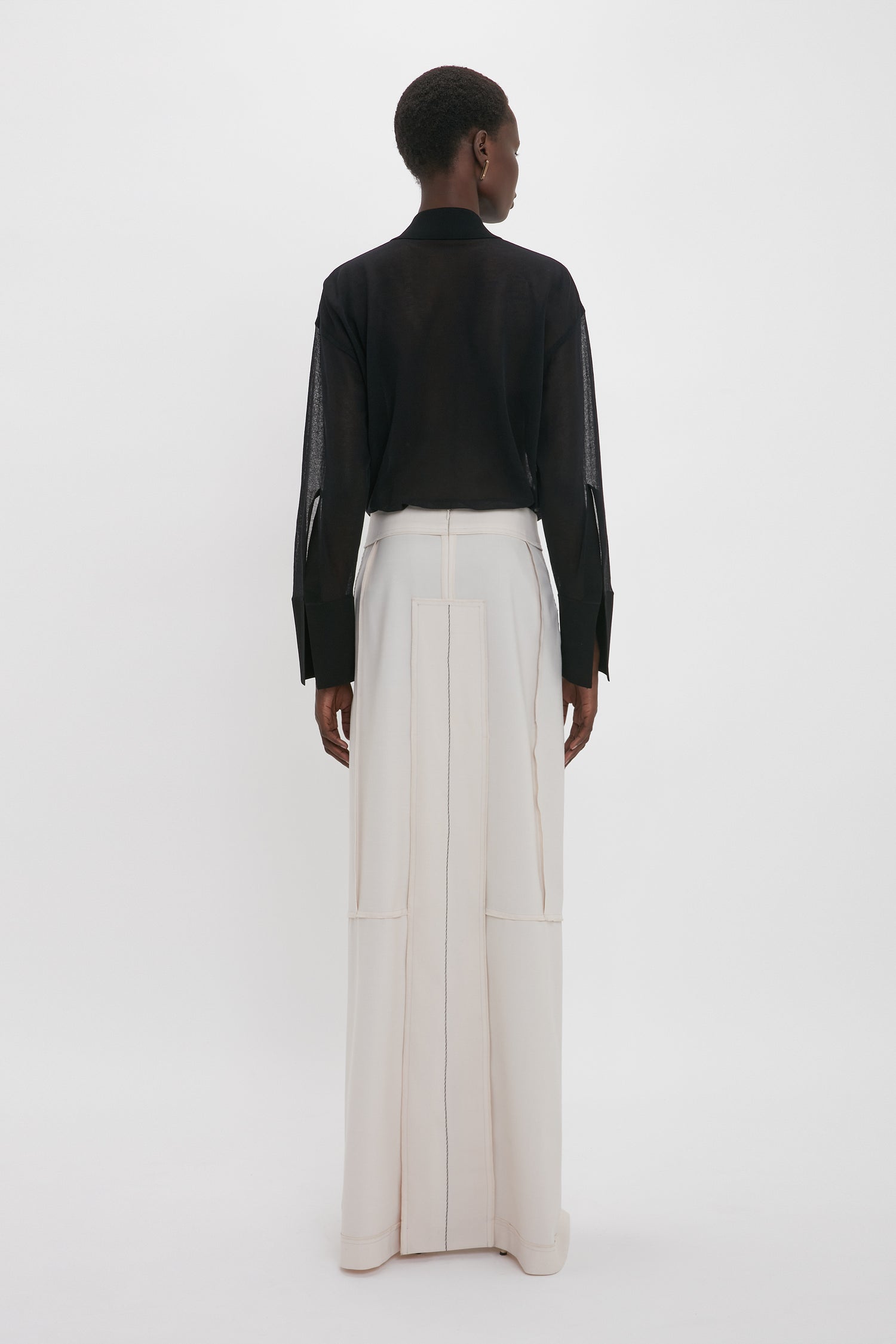 A person stands facing away, wearing a relaxed fit Victoria Beckham Pocket Detail Shirt In Black and high-waisted, wide-leg white trousers with vertical seams.