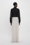 A person stands facing away, wearing a relaxed fit Victoria Beckham Pocket Detail Shirt In Black and high-waisted, wide-leg white trousers with vertical seams.