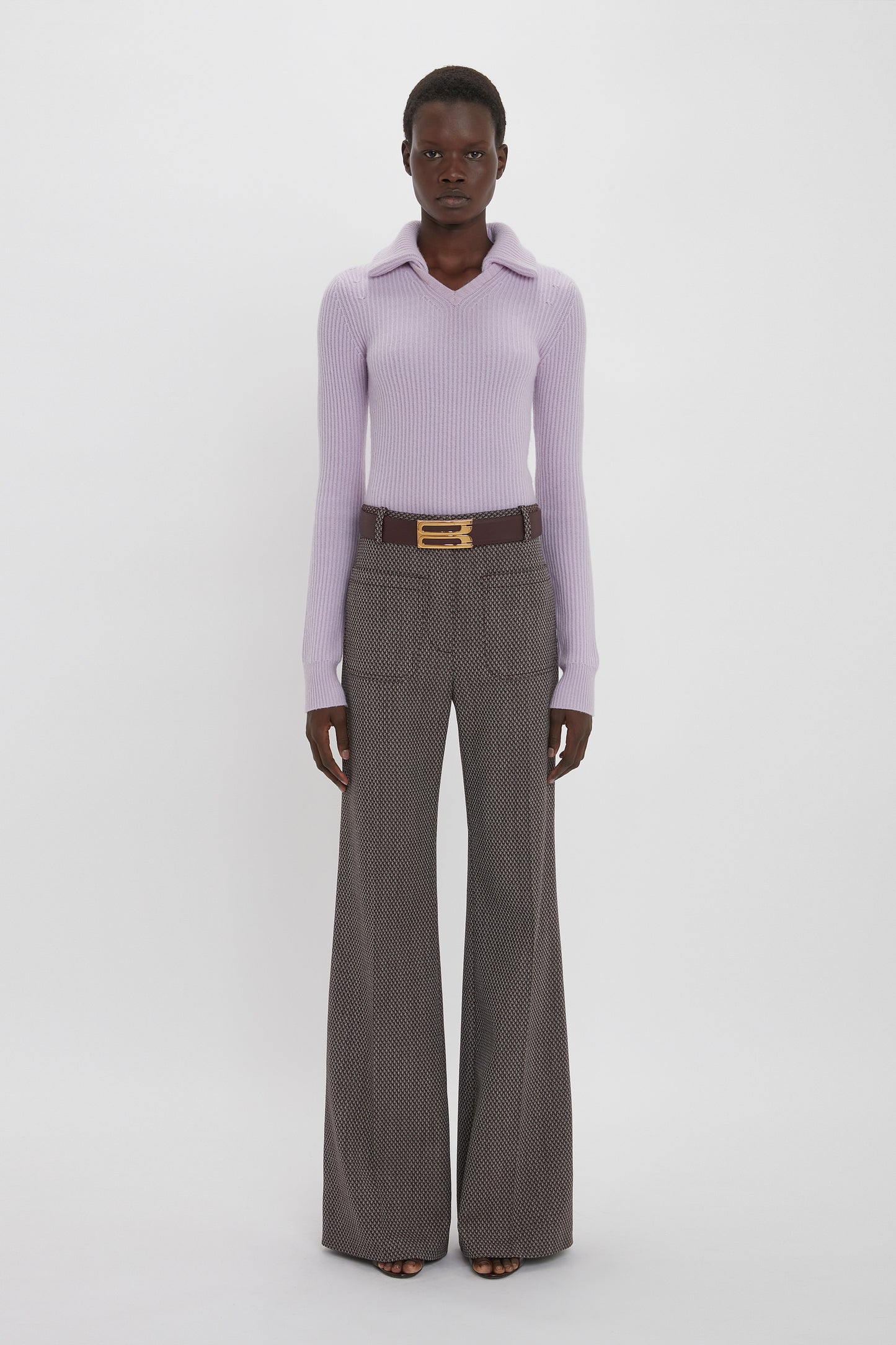 Person stands against a plain background, wearing a light purple ribbed sweater, Alina Trouser In Tobacco by Victoria Beckham with a flared leg and patterned design, and a belt with a gold buckle.