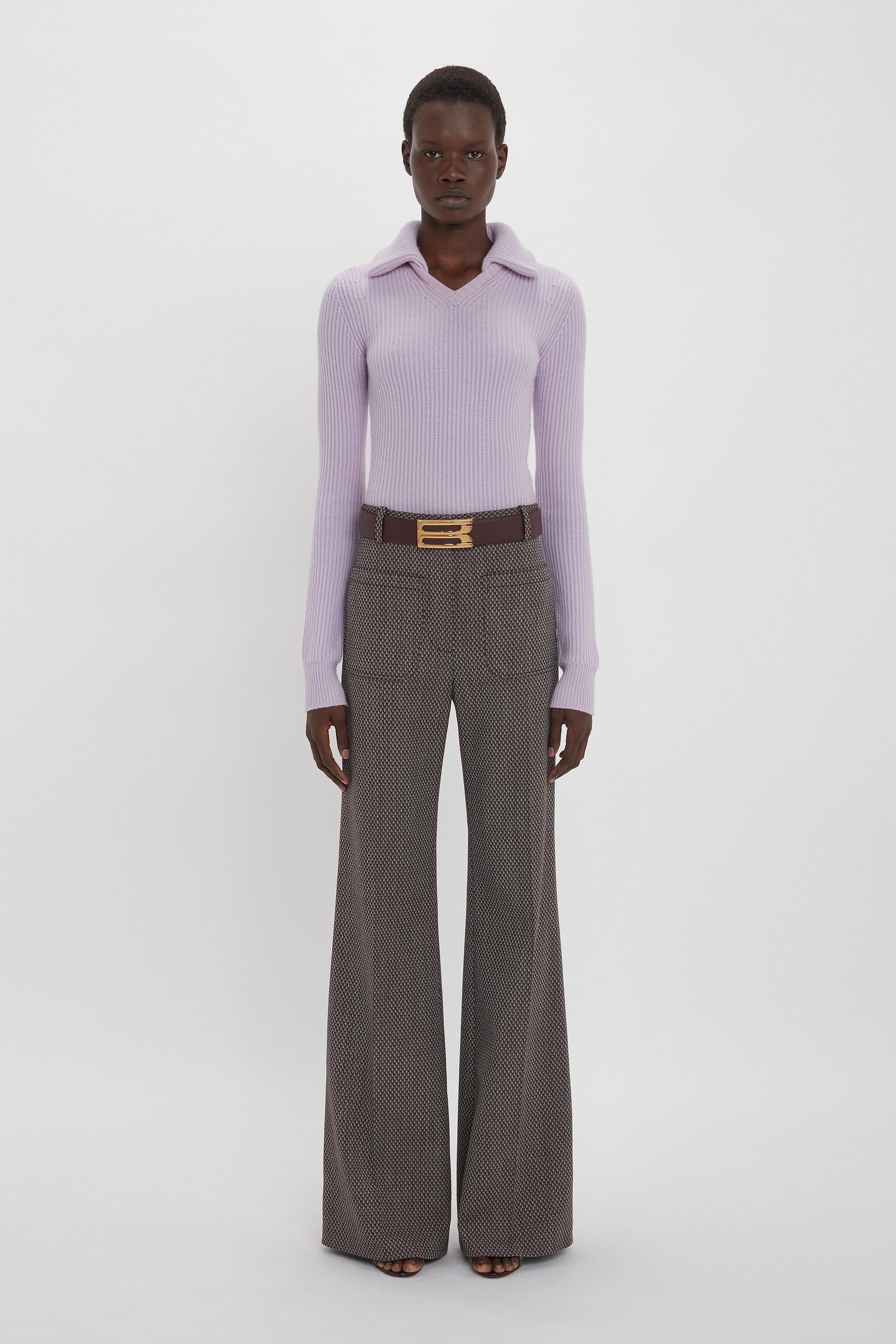 Person stands against a plain background, wearing a light purple ribbed sweater, Alina Trouser In Tobacco by Victoria Beckham with a flared leg and patterned design, and a belt with a gold buckle.