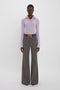 Person stands against a plain background, wearing a light purple ribbed sweater, Alina Trouser In Tobacco by Victoria Beckham with a flared leg and patterned design, and a belt with a gold buckle.