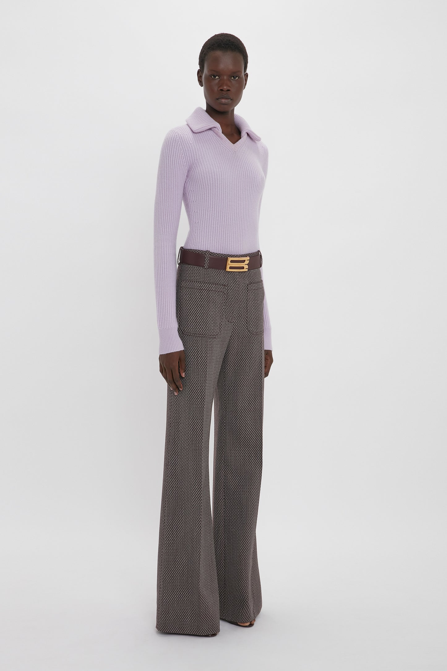 A person stands against a white background, wearing a light purple long-sleeve shirt and Victoria Beckham Alina Trouser In Tobacco with flared legs, finished off with a gold buckle belt.