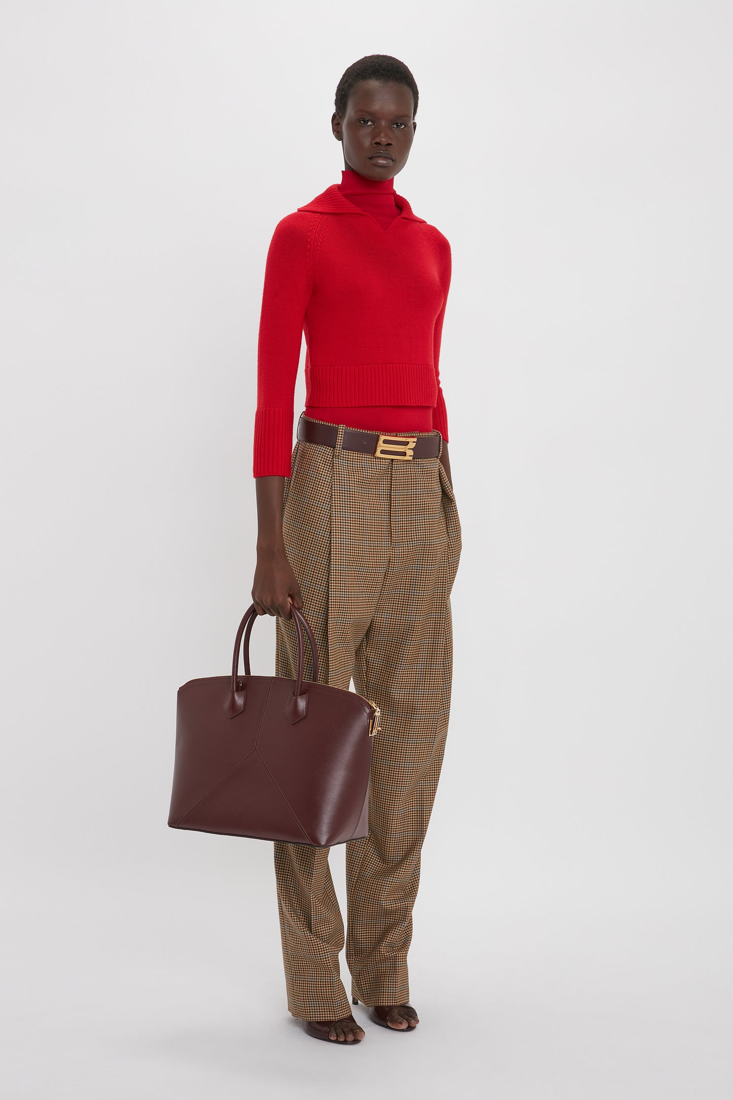 Person standing in a Victoria Beckham Double Layer Top In Deep Red, brown plaid trousers, and holding a large maroon handbag. The person wears a brown belt with a gold buckle. The background is plain white.