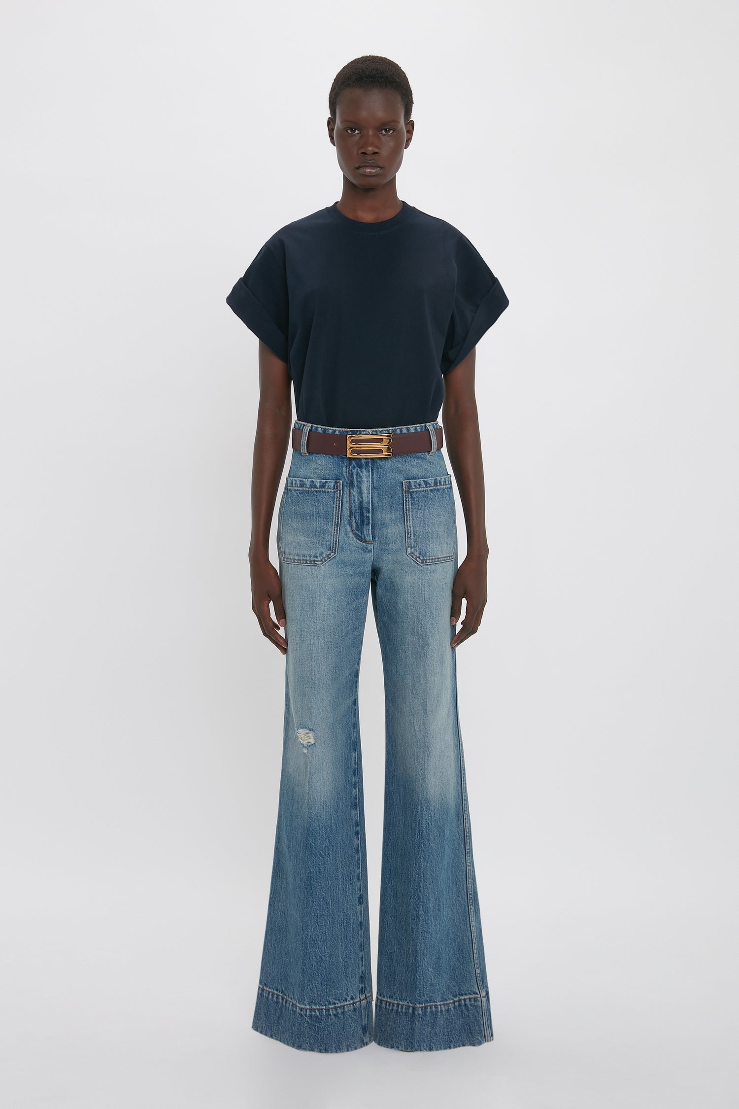 Person standing against a plain white background, wearing a dark short-sleeved Victoria Beckham Asymmetric Relaxed Fit T-Shirt In Navy and high-waisted, wide-leg blue jeans with a brown belt.