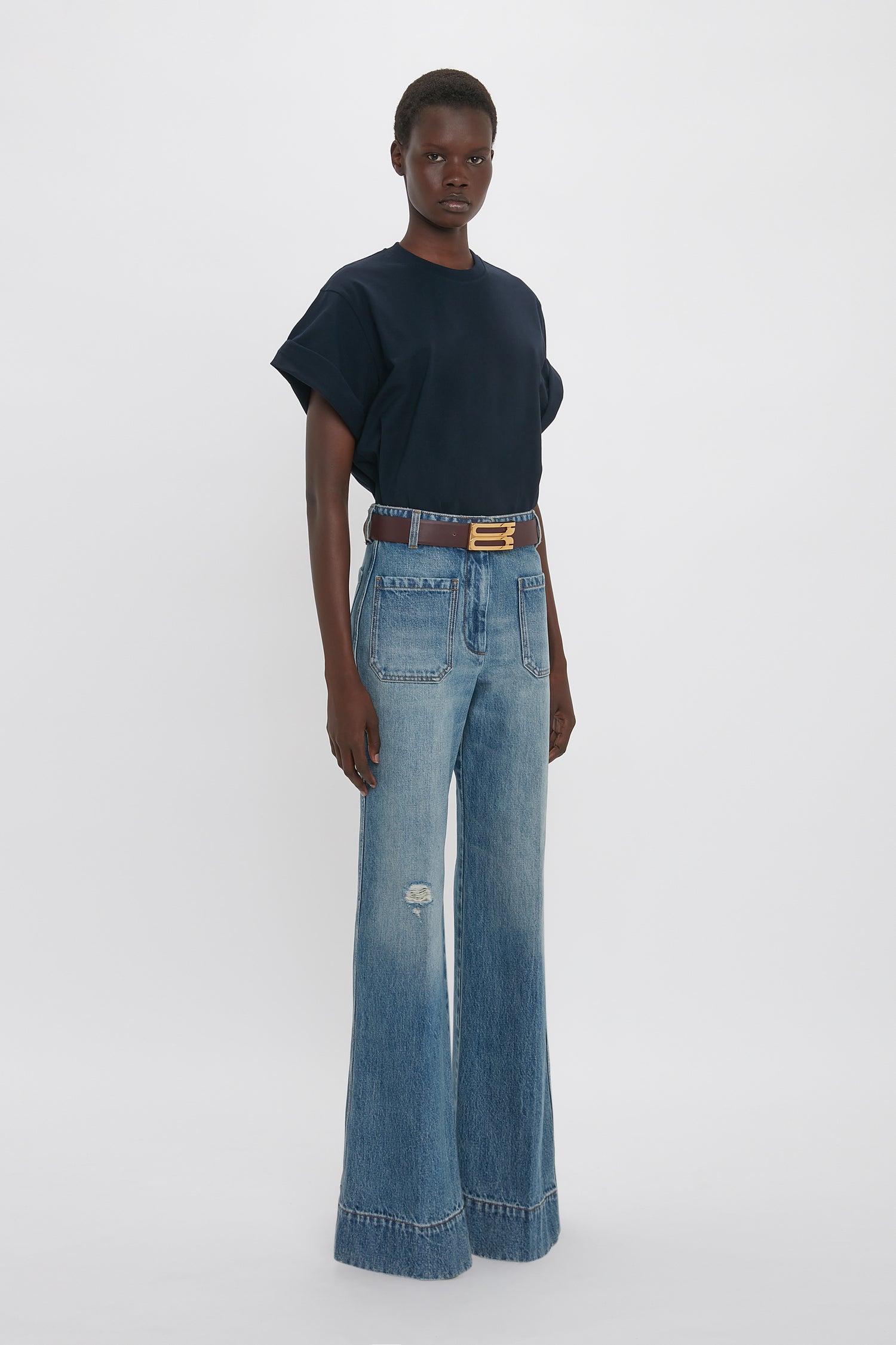 Person wearing an oversized Victoria Beckham Asymmetric Relaxed Fit T-Shirt In Navy with asymmetric folded sleeves, light blue wide-leg jeans with a rip on the left knee, and a brown belt, standing against a plain white background.
