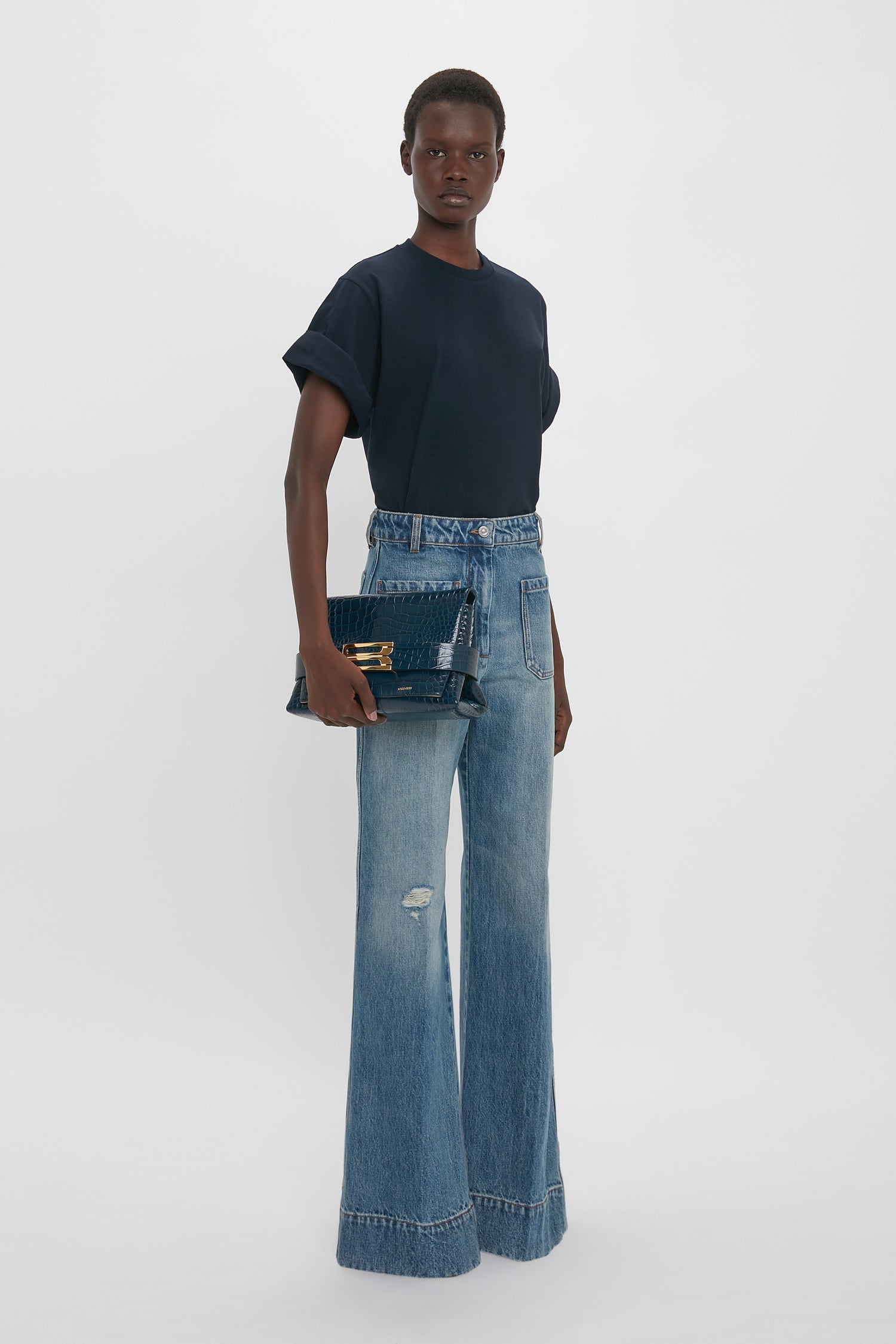 A person stands against a plain background, wearing an oversized navy "Asymmetric Relaxed Fit T-Shirt In Navy" by Victoria Beckham, light blue high-waisted denim pants, and holding a dark blue handbag.
