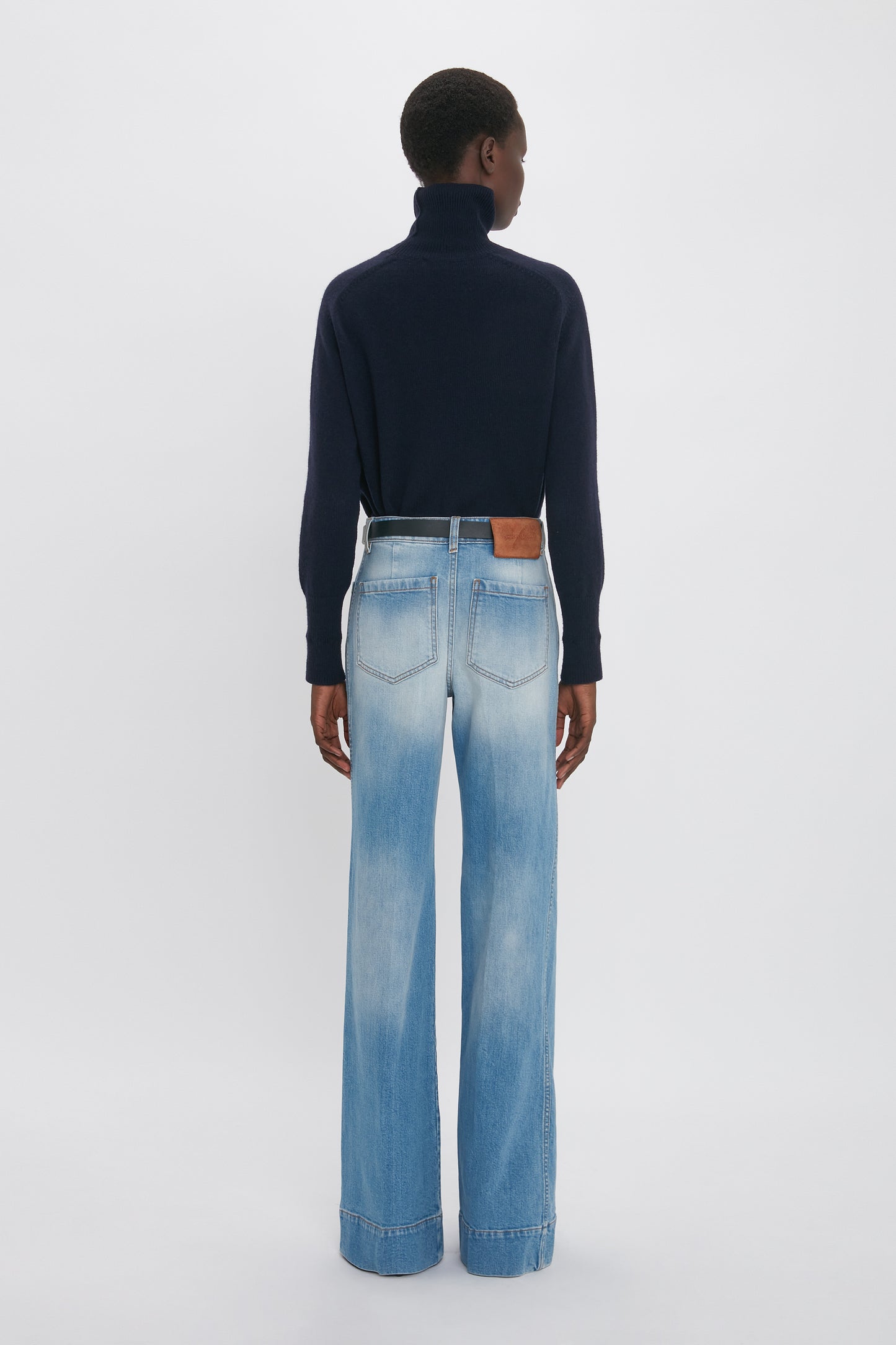 Rear view of a person wearing a dark turtleneck sweater and Victoria Beckham Alina High Waisted Jean In Light Summer Wash against a plain white background.