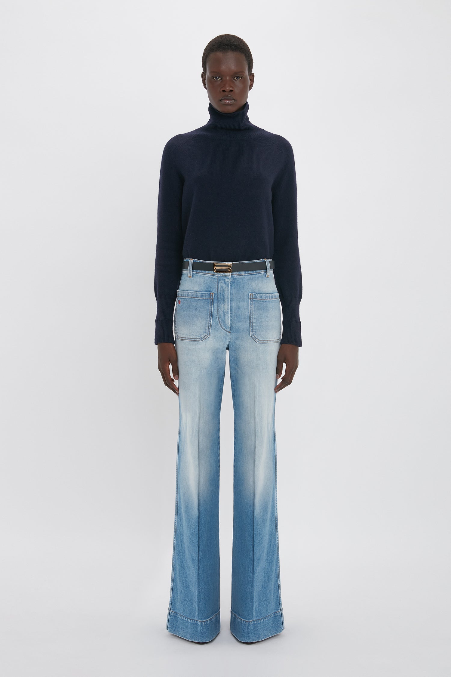 A person stands wearing a dark turtleneck sweater and Victoria Beckham's Alina High Waisted Jean In Light Summer Wash against a plain white background.