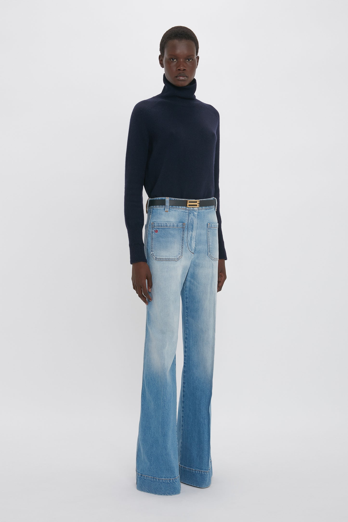 A person stands against a plain background wearing a dark turtleneck sweater and Victoria Beckham's Alina High Waisted Jean In Light Summer Wash with front pockets, showcasing a timeless vintage denim look.