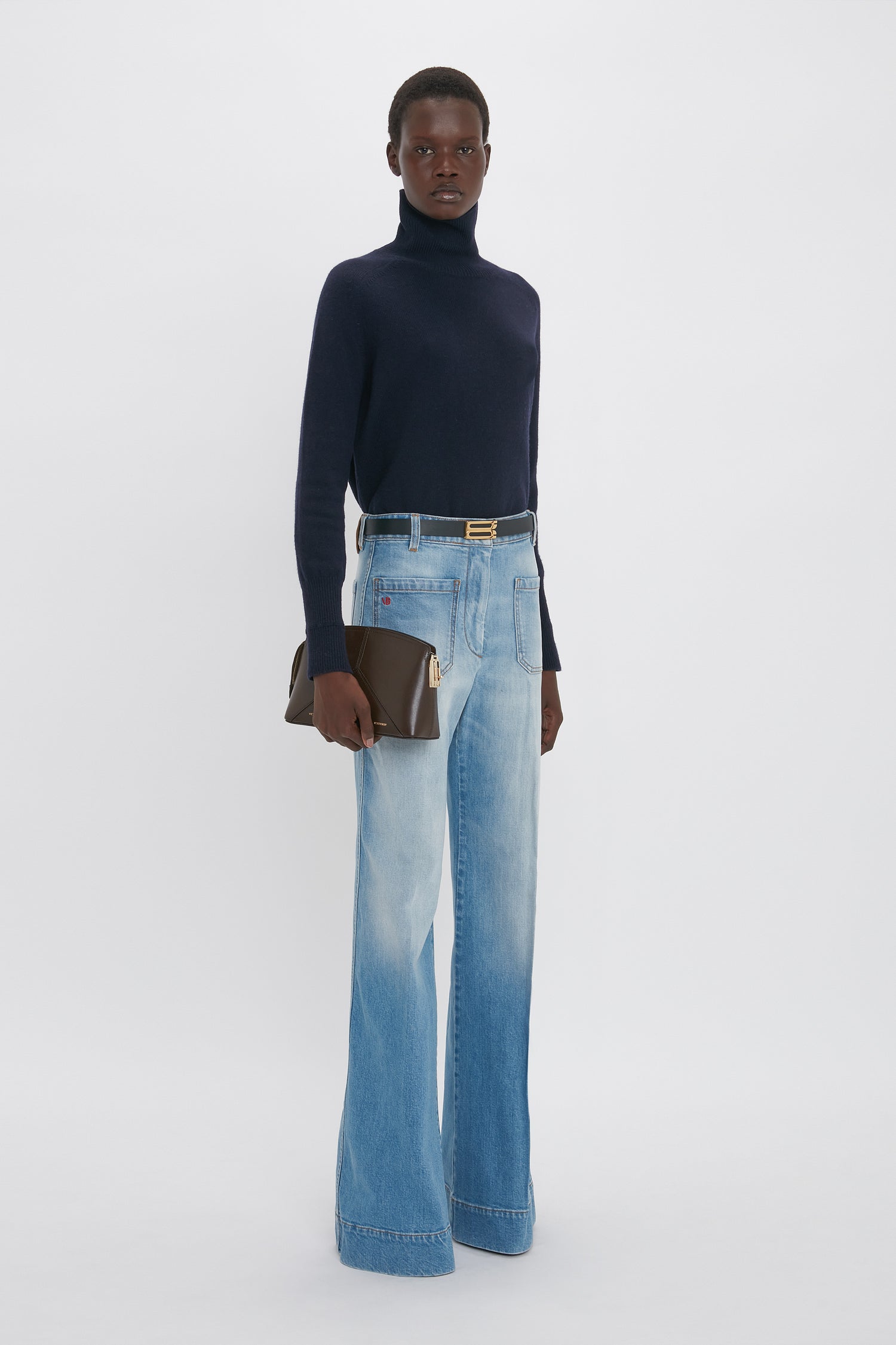 A person stands against a white background, wearing a navy turtleneck, Victoria Beckham Alina High Waisted Jean In Light Summer Wash, and holding a brown clutch bag.