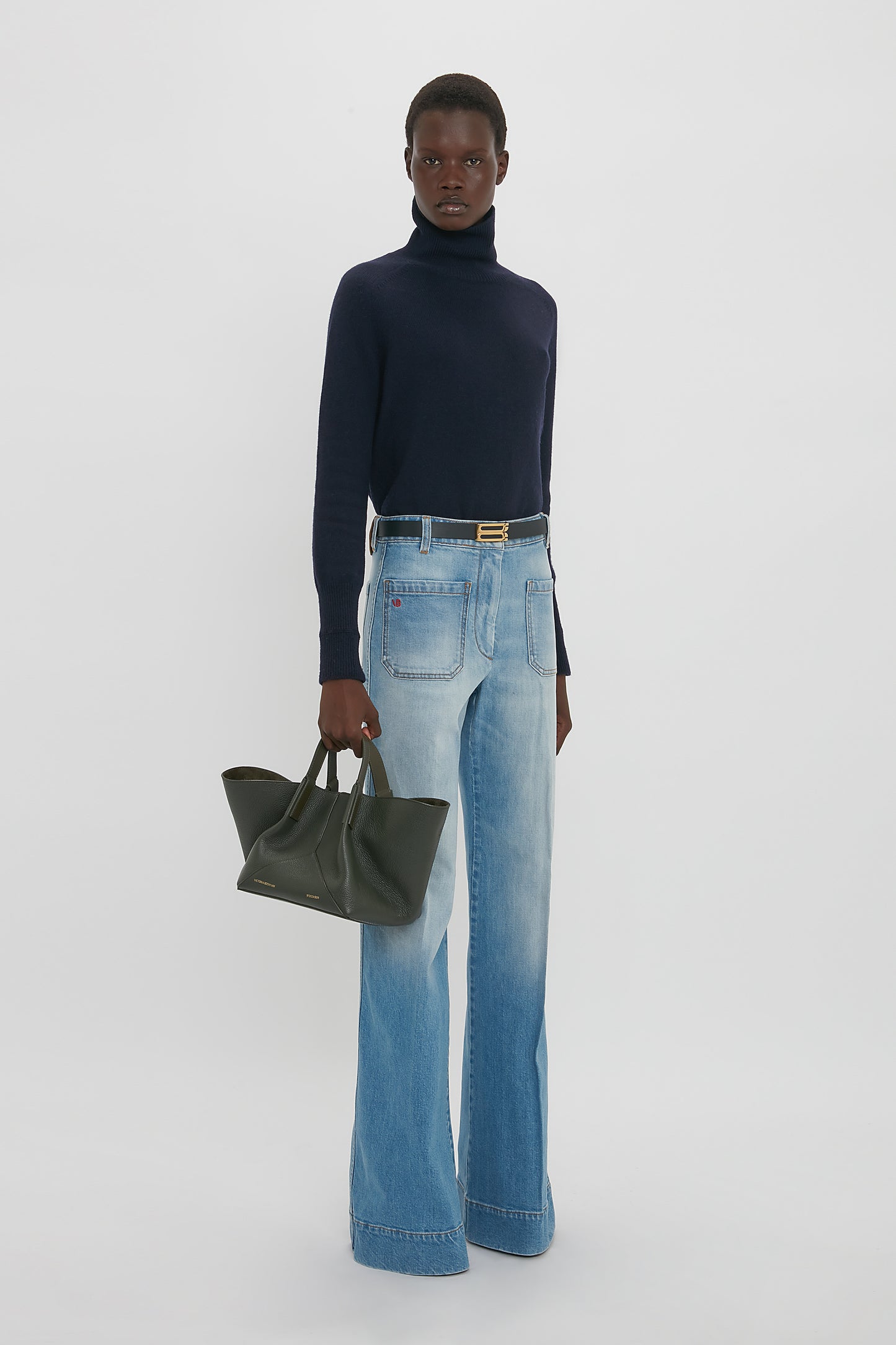 A person stands in a studio wearing a navy turtleneck sweater and light blue wide-leg jeans, holding the Victoria Beckham W11 Mini Tote Bag In Loden Leather. The background is plain white.