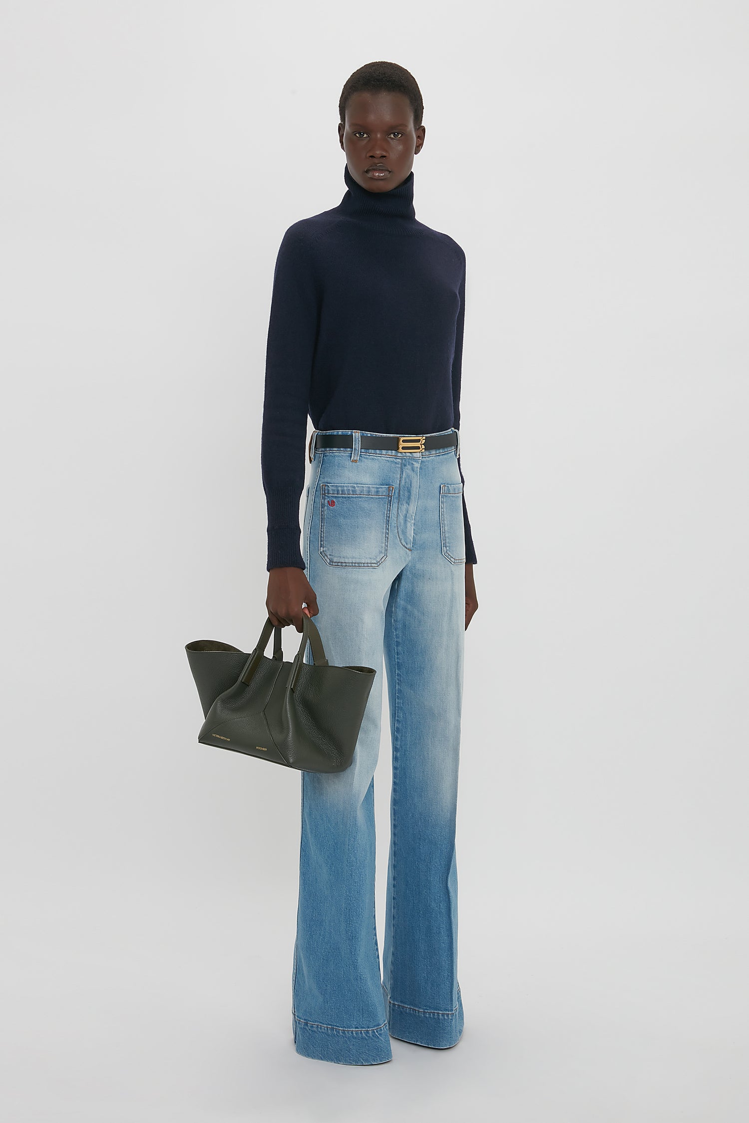 A person stands in a studio wearing a navy turtleneck sweater and light blue wide-leg jeans, holding the Victoria Beckham W11 Mini Tote Bag In Loden Leather. The background is plain white.