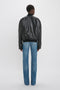 Back view of a person with short hair wearing a Victoria Beckham Leather Varsity Jacket In Black and blue jeans, standing against a plain white background.