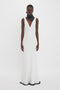 A person wearing a sleeveless white gown with a deep V-neckline and a black scarf stands against a plain white background, flawlessly emulating the sophisticated allure of an Exclusive V-Neck Gathered Waist Floor-Length Gown In Ivory from the Victoria Beckham brand.