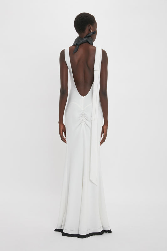 A woman is standing facing away, wearing the Exclusive V-Neck Gathered Waist Floor-Length Gown In Ivory from the Victoria Beckham brand with an open back and a black ribbon tied around her neck. The dress is floor-length with a slight flare at the bottom, showcasing timeless elegance reminiscent of elegant midi dresses.