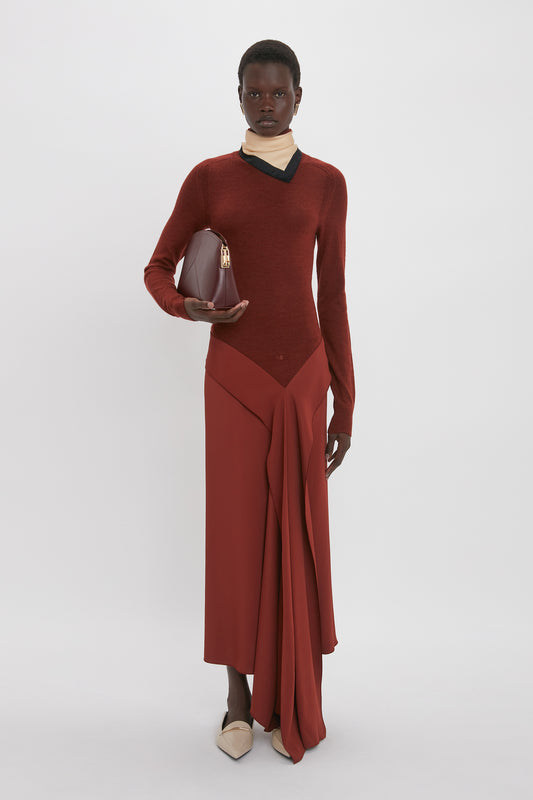A person stands against a plain backdrop, exuding sophistication in a High Neck Tie Detail Dress In Russet by Victoria Beckham. They wear light beige shoes and hold a red handbag, completing the harmonious ensemble with effortless grace.