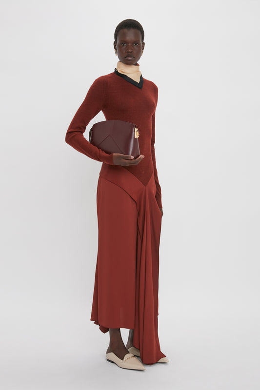 A person stands against a plain background, wearing a red sweater, a red skirt, and beige shoes, holding a red clutch. The ensemble exudes the understated sophistication often associated with Victoria Beckham's High Neck Tie Detail Dress In Russet.