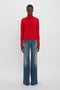 Person wearing a red turtleneck sweater and Victoria Beckham Alina High Waisted Jean In Heavy Vintage Indigo Wash stands against a plain white background.