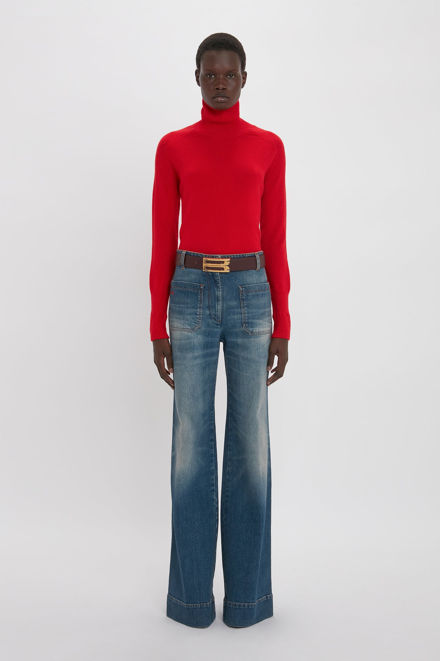 Person wearing a red Victoria Beckham Polo Neck Jumper In Red and wide-leg blue jeans standing against a plain white background.