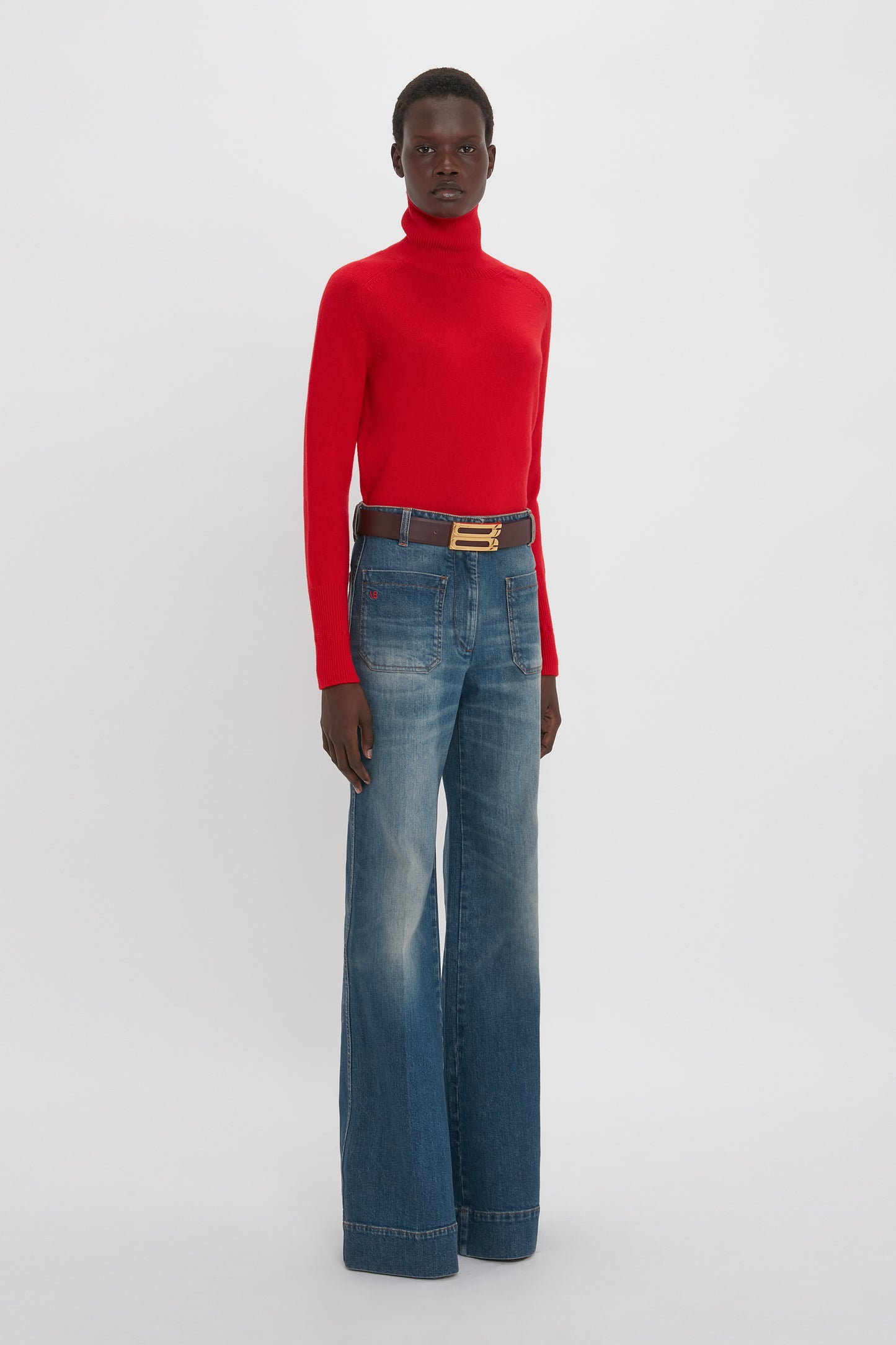 A person stands against a white background wearing a Polo Neck Jumper In Red by Victoria Beckham, blue wide-leg jeans, and a black belt with a large buckle.