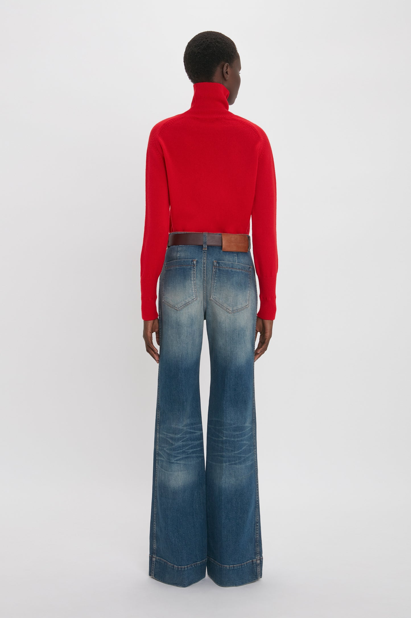 Person wearing a red Victoria Beckham Polo Neck Jumper In Red and high-waisted, wide-leg blue jeans, standing with their back facing the camera against a plain white background.