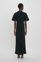 A person wearing a Gathered V-Neck Midi Dress in Black by Victoria Beckham stands with their back facing the camera against a plain white background, exemplifying contemporary edge. The figure-flattering stretch fabric subtly enhances their silhouette.