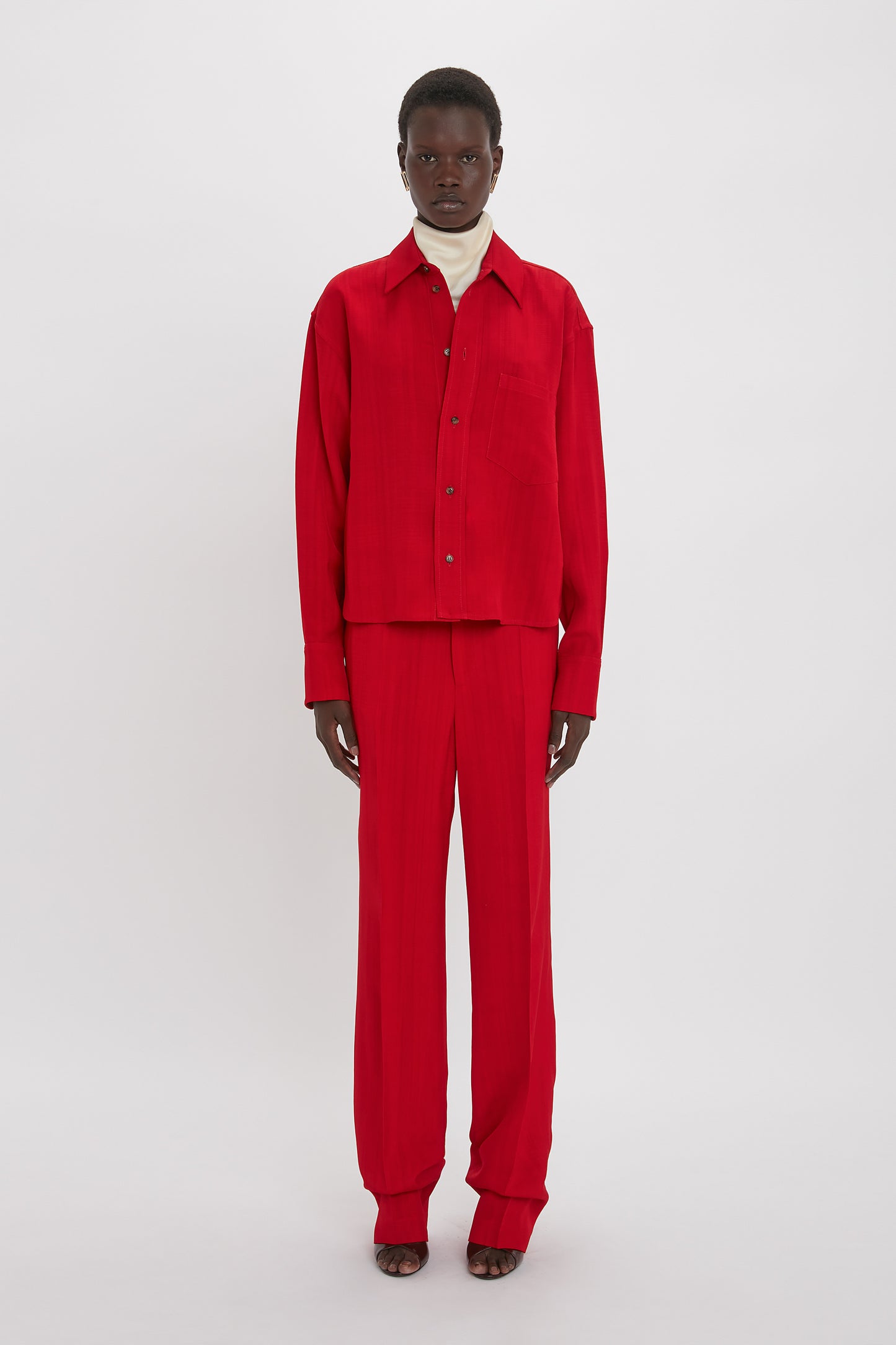 A person stands facing forward, wearing a red Cropped Long Sleeve Shirt In Carmine by Victoria Beckham with a masculine-inspired design, paired with matching red pants and a white turtleneck underneath.
