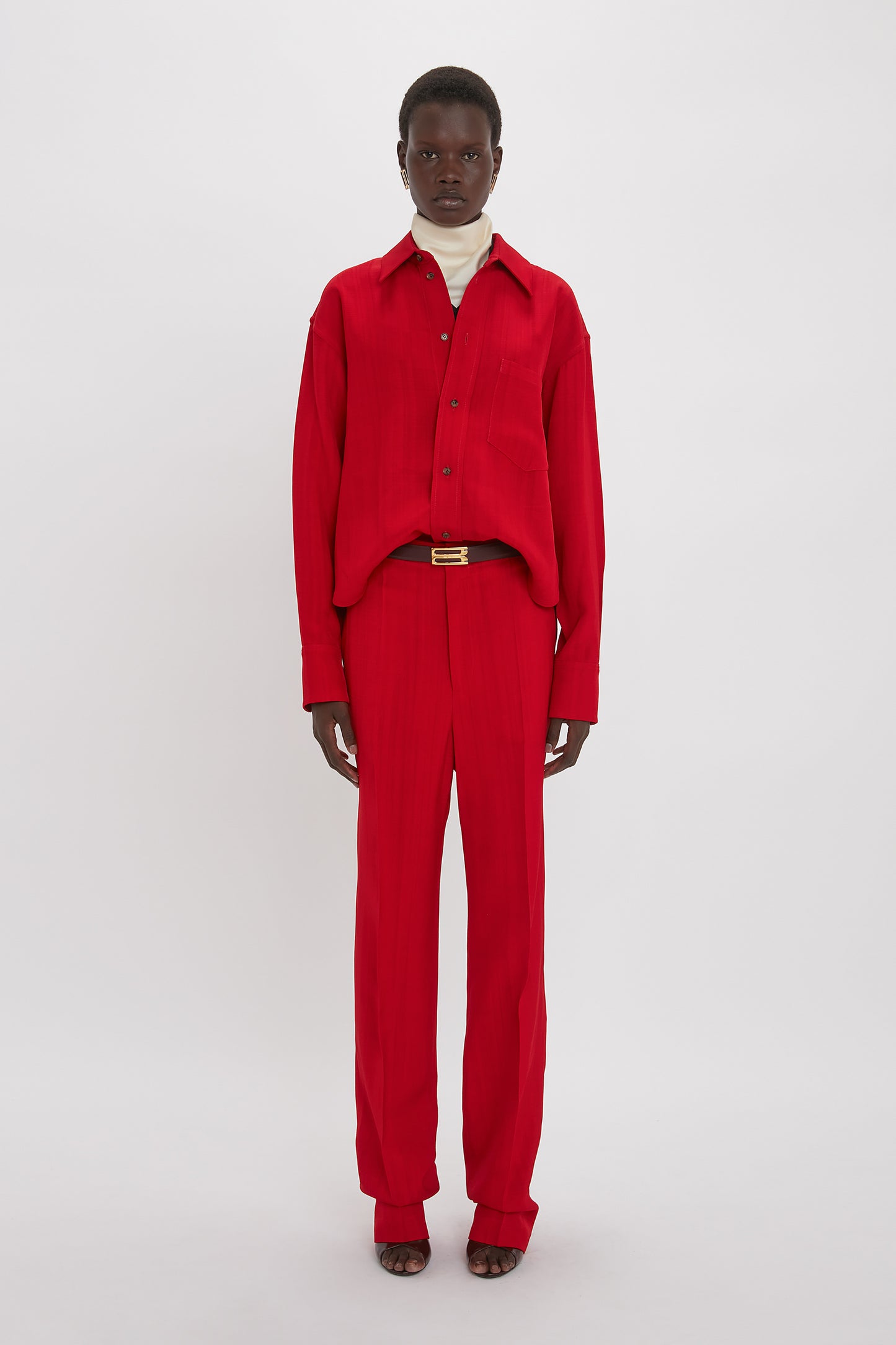 A person stands against a plain backdrop wearing a Victoria Beckham Cropped Long Sleeve Shirt In Carmine and matching red pants, with a white turtleneck shirt visible underneath. The ensemble features a masculine-inspired shirt design that adds an edge to the sophisticated look.