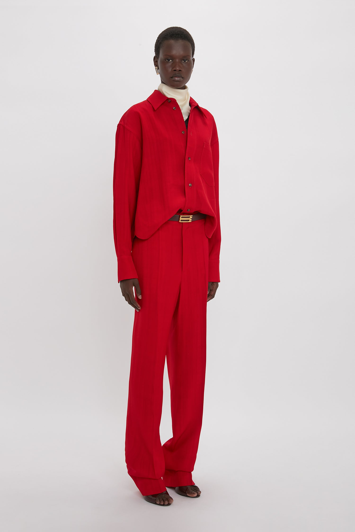 A person stands against a plain background, wearing the Cropped Long Sleeve Shirt In Carmine by Victoria Beckham, matching red pants, and black sandals.