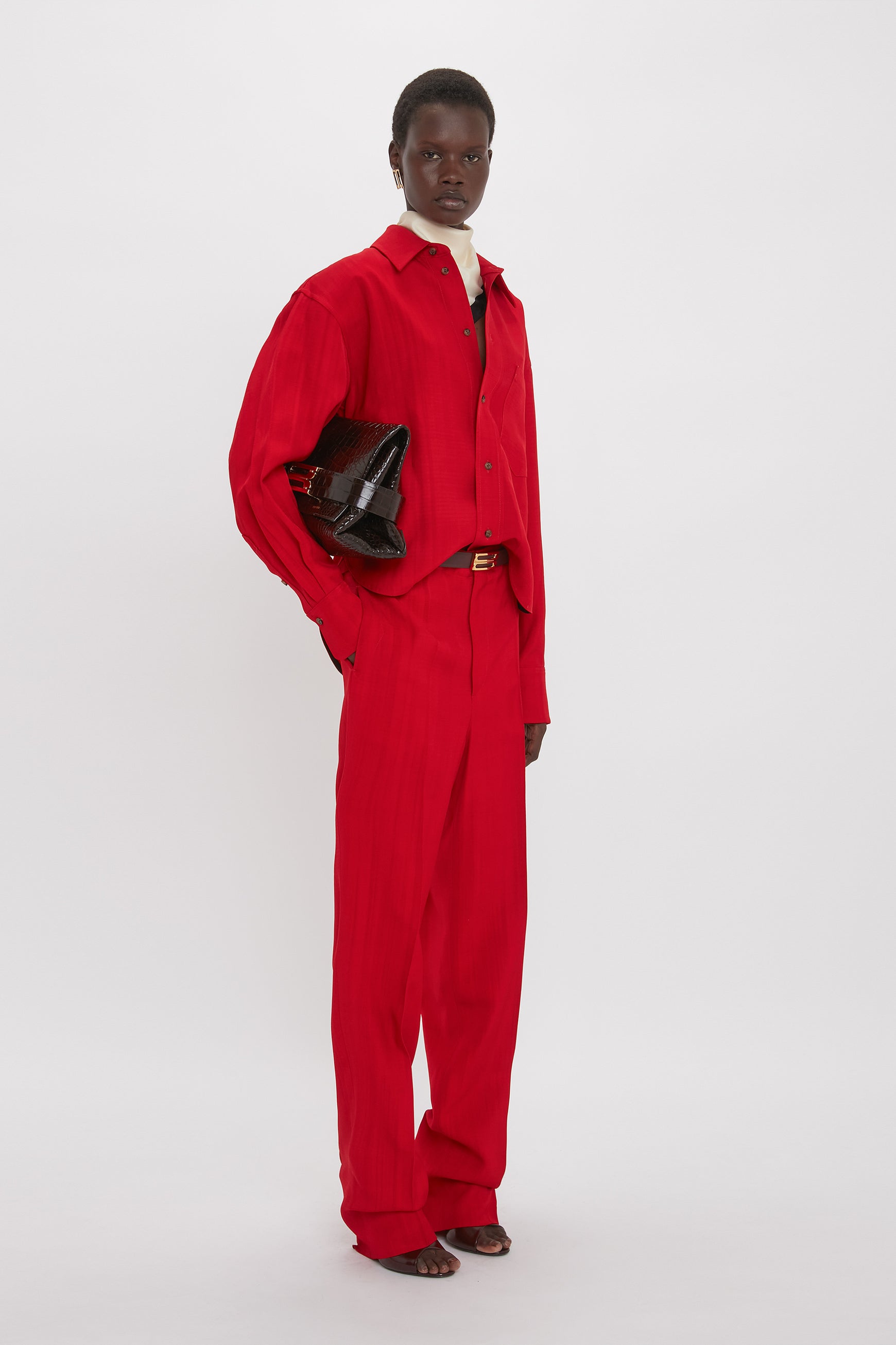 Person standing against a plain backdrop, wearing matching red pants and shirt, holding a B Pouch Bag In Croc Effect Espresso Leather by Victoria Beckham in their left hand, and wearing sandals.
