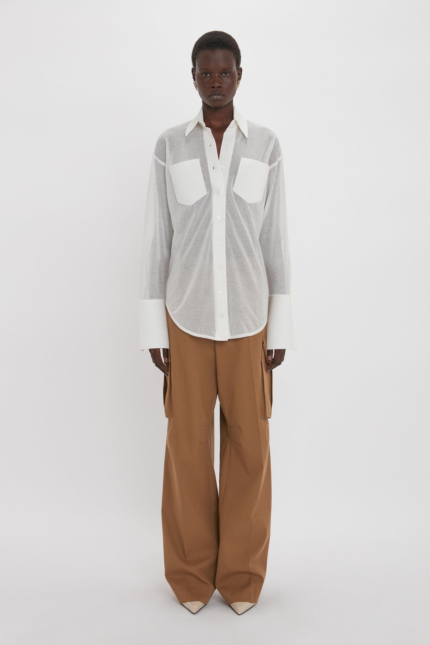 Person standing against a plain background wearing a sheer Pocket Detail Shirt In White by Victoria Beckham over a white top, paired with tan high-waisted wide-leg pants, exemplifying casual sophistication with its relaxed fit and large chest pockets.