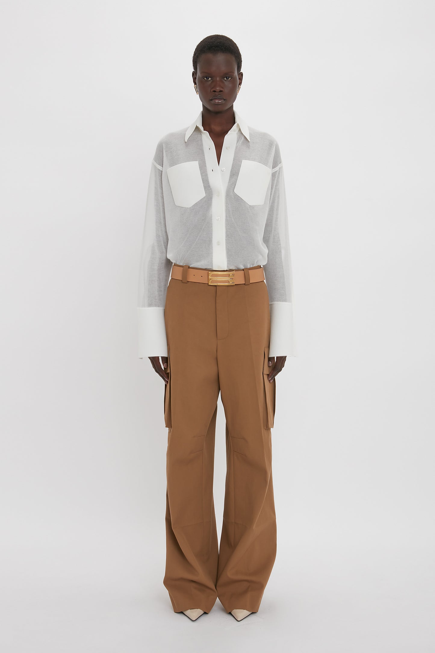 Person standing against a plain white background, wearing the Victoria Beckham Pocket Detail Shirt In White with prominent pockets and brown wide-legged pants, secured with a belted waist—an ensemble radiating casual sophistication.