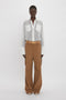 Person standing against a plain white background, wearing the Victoria Beckham Pocket Detail Shirt In White with prominent pockets and brown wide-legged pants, secured with a belted waist—an ensemble radiating casual sophistication.