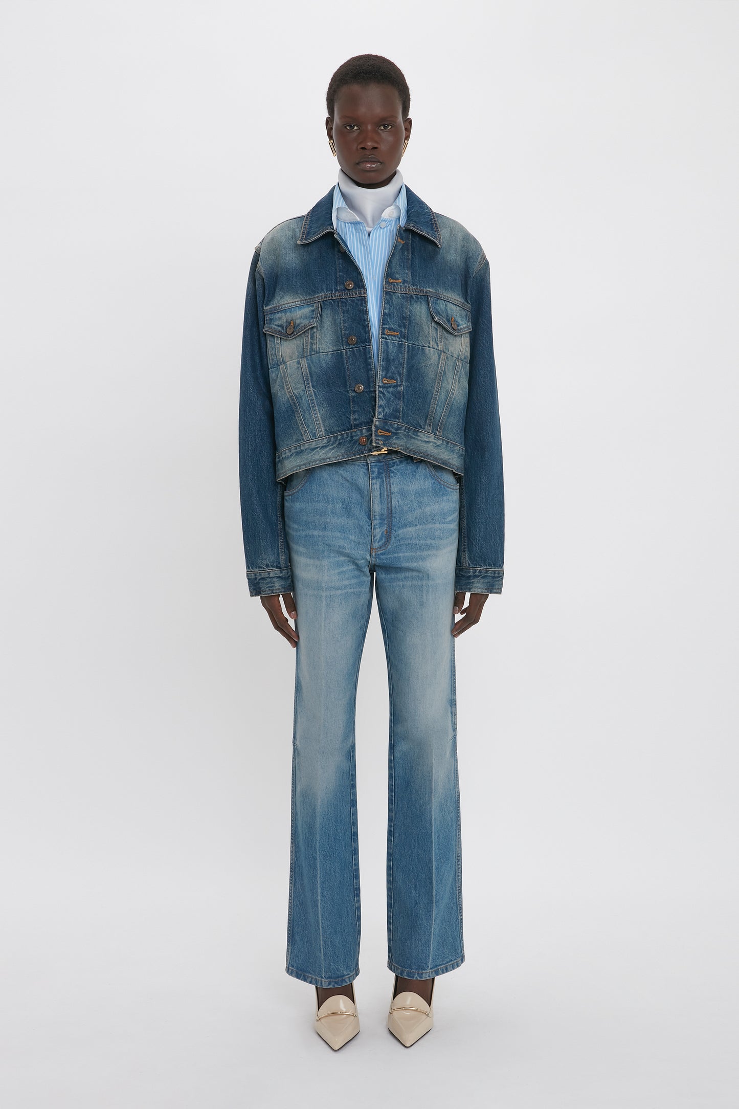 Person standing against a plain background, wearing a Cropped Denim Jacket In Heavy Vintage Indigo Wash by Victoria Beckham over a striped shirt, paired with vintage wash faded jeans and beige pointed toe shoes.