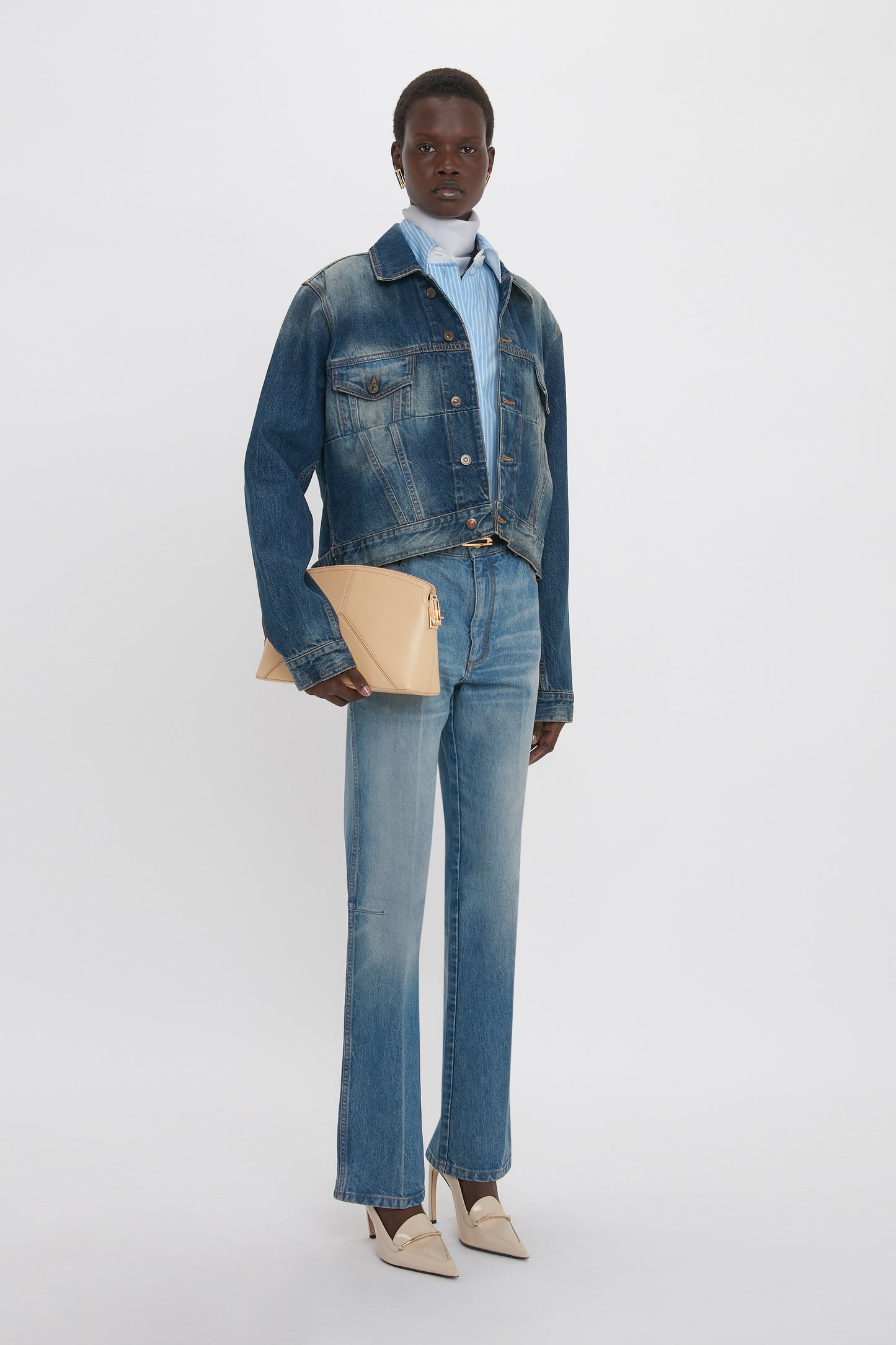 Person standing against a plain background, dressed in a Cropped Denim Jacket In Heavy Vintage Indigo Wash by Victoria Beckham and jeans with a vintage wash, holding a beige clutch bag. They are wearing a light blue shirt and beige heels, showcasing a stylishly feminine silhouette.