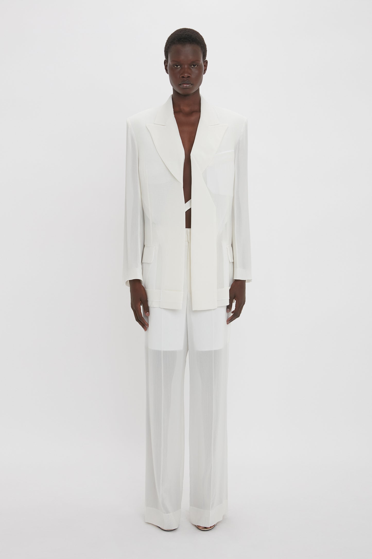 A person is standing against a white background, wearing the Victoria Beckham Fold Detail Tailored Jacket In White with matching wide-leg pants.