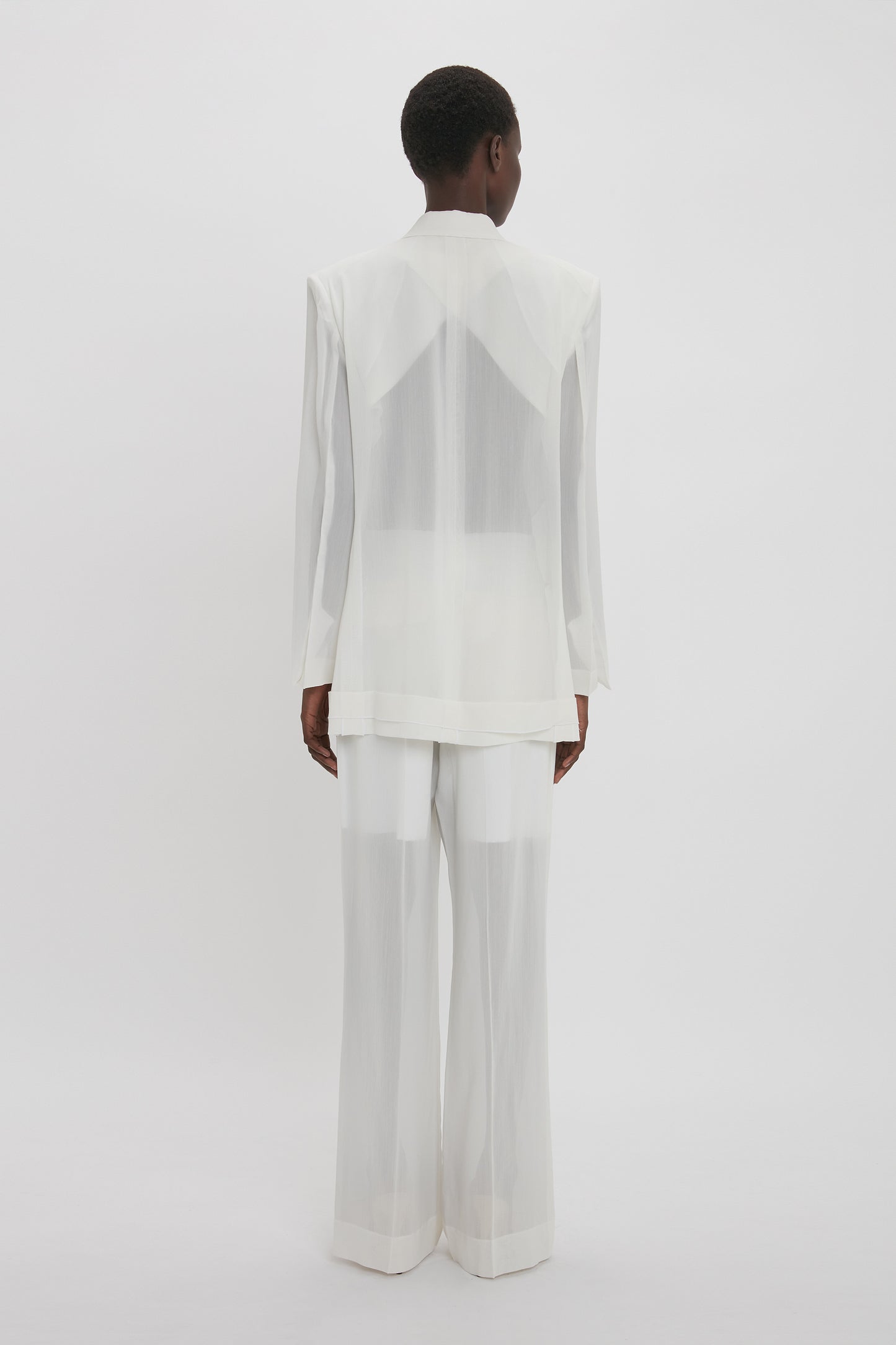 A person is standing facing away from the camera, wearing a Fold Detail Tailored Jacket In White by Victoria Beckham with wide-legged pants against a plain white background.