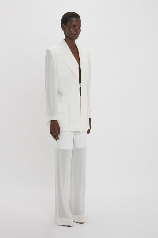A person is standing against a plain backdrop, dressed in the Victoria Beckham Fold Detail Tailored Jacket In White with wide lapels and matching wide-legged pants that feature a chic satin collar.