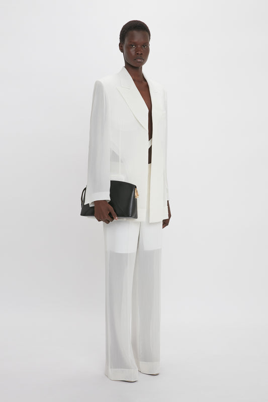 A person stands against a plain backdrop, wearing a white suit featuring the Fold Detail Tailored Jacket In White by Victoria Beckham with a double-breasted cut and wide-legged pants, holding a black clutch bag.